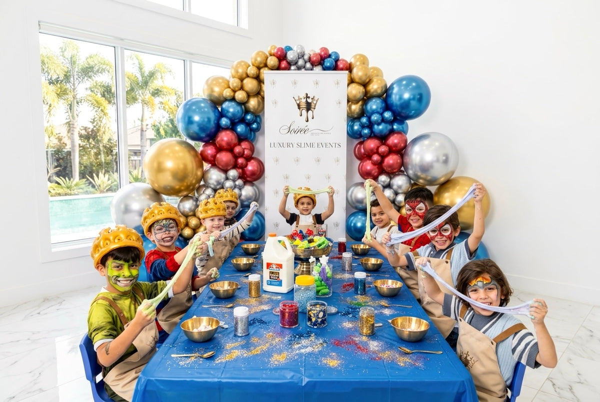 Children in gold crowns and superhero face paint stretching slime along a blue party table under a balloon arch